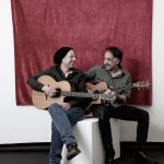 Zwei Männer mit Gitarre sitzen einander zugewandt und lächeln