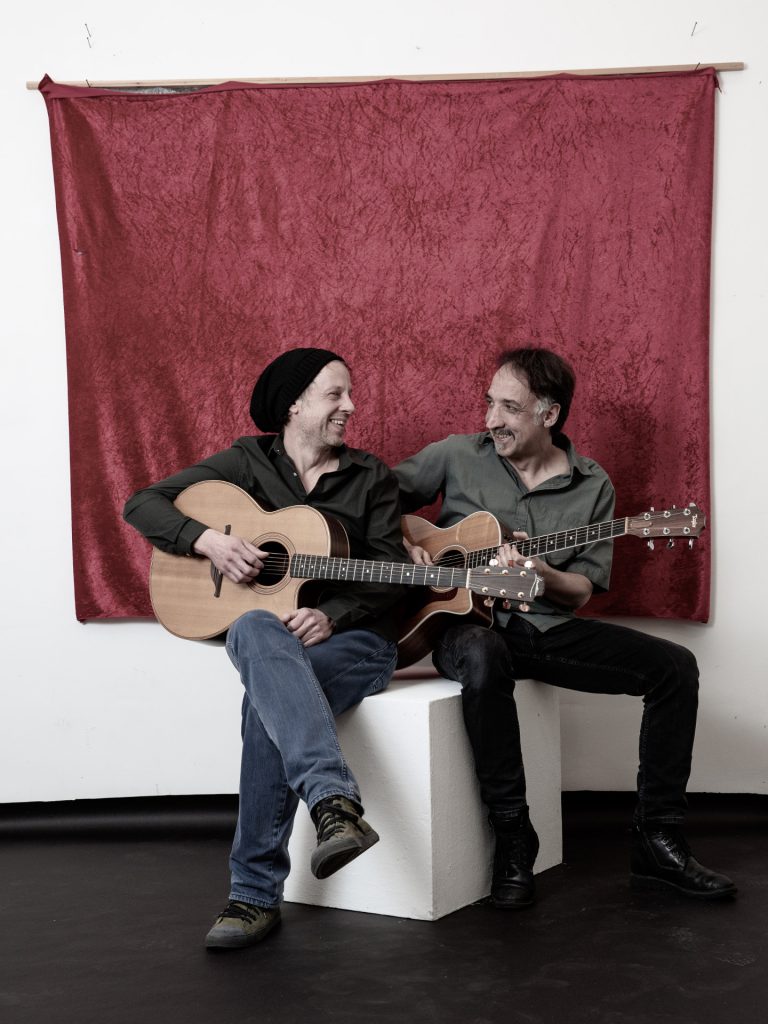 Zwei Männer mit Gitarre sitzen einander zugewandt und lächeln