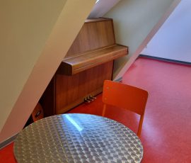 In einem Dachraum stehen ein Klavier und ein Tisch mit Stühlen